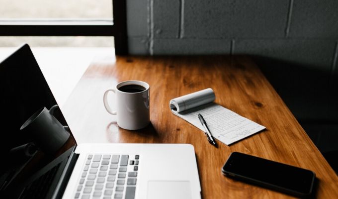 baner przedstawia stół, na którym leży laptop, smartfon, notes wraz z długopisem oraz kubek kawy